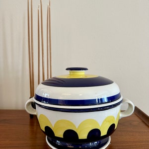 May include: A white ceramic serving bowl with a blue and yellow geometric pattern. The bowl has a lid and a base. The bowl is on a wooden surface.