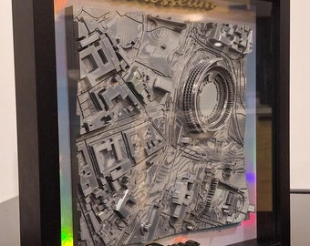 Può includere: Un'opera d'arte tridimensionale incorniciata raffigurante il Colosseo a Roma. La mappa in rilievo grigia è racchiusa in una cornice nera, con la parola "Colosseum" in oro. L'opera d'arte è esposta sopra un pezzo simile, con il logo "LOCK3DIN" visibile.