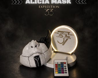 May include: A white and black mask with geometric patterns, accompanied by a circular illuminated display stand featuring the text FOR THOSE WHO COME AFTER 33. A multi-coloured remote control is positioned in front of the display stand.
