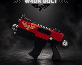Può includere: Un'arma di scena rossa e nera con un emblema dorato a forma di aquila bicipite sul lato. Il testo W40K BOLT appare in alto, con THE IMPERIUM OF MAN e LOCK3DIN stampati in basso. Il design include un caricatore e un'impugnatura neri.