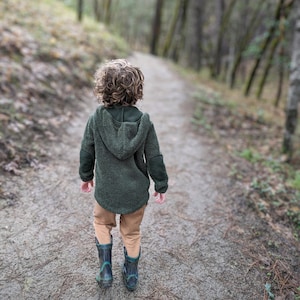 Puede incluir: Un ni&ntilde;o con una chaqueta con capucha verde oscuro, pantalones color canela y botas de lluvia azul oscuro camina por un sendero forestal. La chaqueta tiene un aspecto texturizado y difuso. El sendero est&aacute; rodeado de &aacute;rboles.