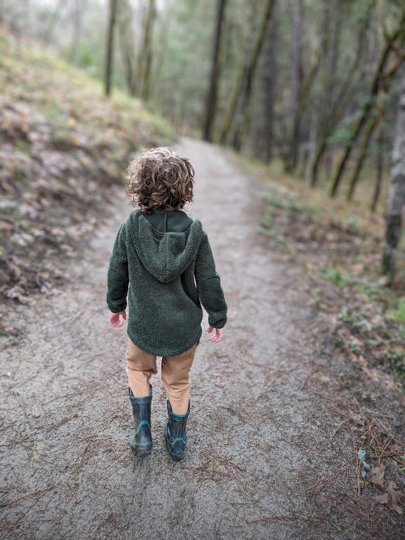 Puede incluir: Un ni&ntilde;o con una chaqueta con capucha verde oscuro, pantalones color canela y botas de lluvia azul oscuro camina por un sendero forestal. La chaqueta tiene un aspecto texturizado y difuso. El sendero est&aacute; rodeado de &aacute;rboles.