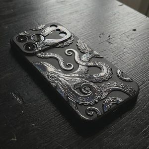 May include: A matte black smartphone case featuring a raised, metallic silver octopus design that wraps around the camera module. The intricate tentacles with detailed suction cups extend across the back surface, creating a textured, three-dimensional effect.