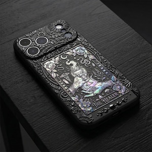 May include: A black phone case featuring an intricate, embossed design with iridescent accents. The central graphic depicts two hands holding a chalice, surrounded by floral motifs, snakes, and the text THE HEALER. The surface is textured with celestial symbols.