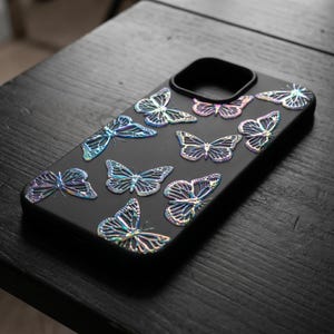 May include: Black phone case adorned with iridescent butterfly embellishments. The butterflies are in various sizes and orientations, creating a decorative pattern. The case has a sleek, modern design.