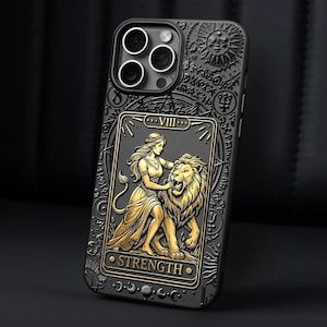K&ouml;nnte beinhalten: Schwarze Handyh&uuml;lle mit einem goldenen Design der Tarotkarte St&auml;rke. Die H&uuml;lle zeigt eine Frau mit einem L&ouml;wen, darunter das Wort &bdquo;STRENGTH&ldquo;. Die H&uuml;lle hat eine strukturierte Oberfl&auml;che und einen Kameraausschnitt.