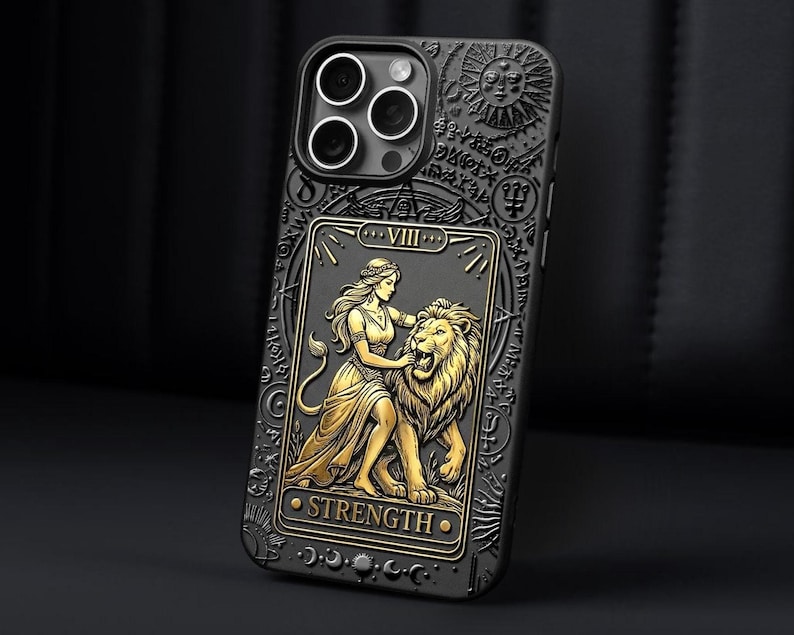 K&ouml;nnte beinhalten: Schwarze Handyh&uuml;lle mit einem goldenen Design der Tarotkarte St&auml;rke. Die H&uuml;lle zeigt eine Frau mit einem L&ouml;wen, darunter das Wort &bdquo;STRENGTH&ldquo;. Die H&uuml;lle hat eine strukturierte Oberfl&auml;che und einen Kameraausschnitt.