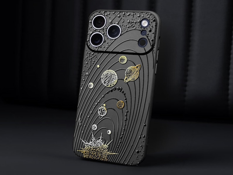 Peut inclure: Coque de smartphone noire avec un motif plan&eacute;taire d&eacute;taill&eacute;. L'&eacute;tui pr&eacute;sente une surface textur&eacute;e avec des lignes en relief et des accents m&eacute;talliques dor&eacute;s et argent&eacute;s repr&eacute;sentant des plan&egrave;tes et des &eacute;toiles. Les objectifs de l'appareil photo sont visibles en haut.