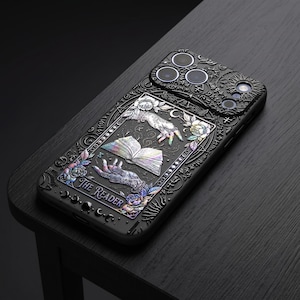 May include: Black phone case with an intricate embossed design. The case features a tarot card theme with hands, an open book, and floral accents. The text "The Reader" is visible. The case has a camera lens protector.