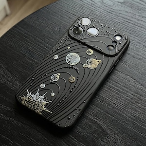 Peut inclure: Coque de t&eacute;l&eacute;phone noire avec un motif de syst&egrave;me solaire en relief. La coque pr&eacute;sente des plan&egrave;tes, des &eacute;toiles et un motif de soleil en or et argent m&eacute;talliques. La coque a une surface textur&eacute;e.