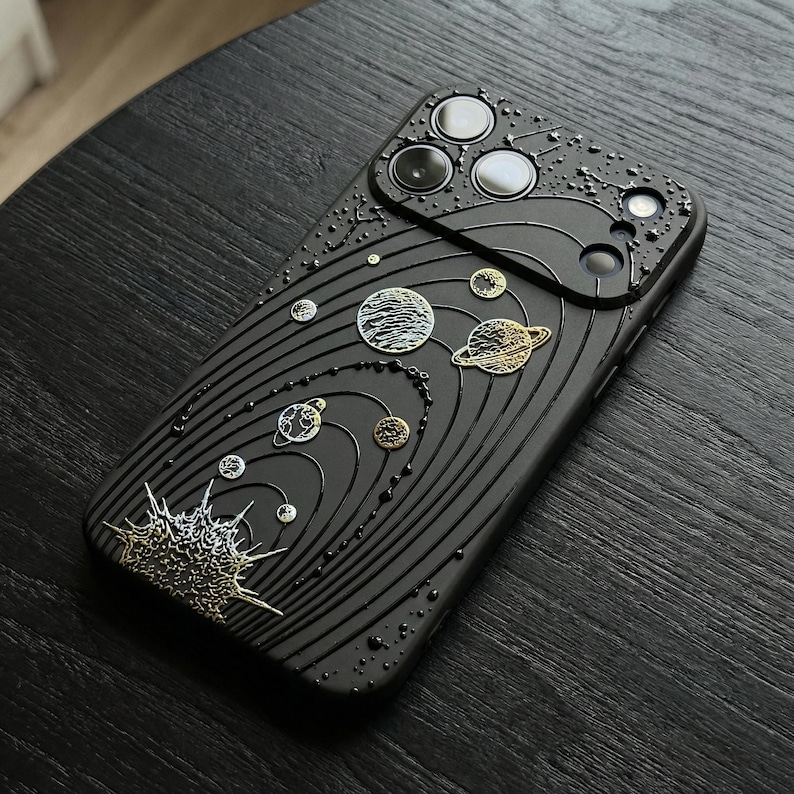 Peut inclure: Coque de t&eacute;l&eacute;phone noire avec un motif de syst&egrave;me solaire en relief. La coque pr&eacute;sente des plan&egrave;tes, des &eacute;toiles et un motif de soleil en or et argent m&eacute;talliques. La coque a une surface textur&eacute;e.