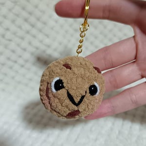 Peut inclure: Un porte-clés cookie crocheté, rond et beige, avec une texture en relief, des pépites de chocolat marron foncé et un visage brodé composé d'yeux noirs et d'une bouche en forme de V. Le cookie en peluche est fixé à un anneau et une chaîne dorés.