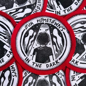 May include: A collection of circular patches with a red border and white stitching. Each patch features a black and white illustration with the text "OUR HOMETOWN'S IN THE DARK". The design includes a figure with a circular head and abstract shapes.