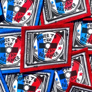 May include: A square fabric patch with a red border featuring a circular graphic design. The center contains the text "POWER TO THE LOCAL DREAMER" in black, overlaid on a blue and red diagonal background with small blue and red stars.