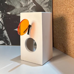 May include: A white rectangular clock with a circular orange accent and black hands. The clock has a circular opening and is set against a black and white abstract painting and a corkboard.