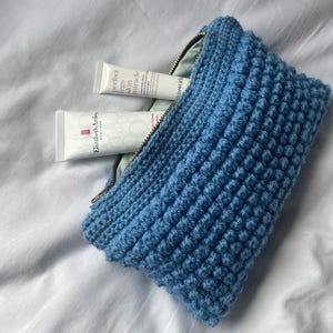 May include: A blue knitted cosmetic bag with a zipper, containing two white tubes of skincare products. The bag has a textured, bobble stitch design. The zipper is gold-colored.