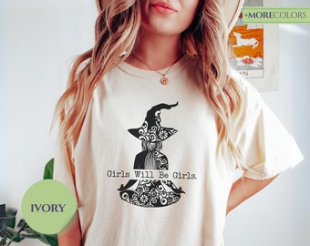 May include: Ivory-colored t-shirt featuring a black graphic of a witch in a hat, with the text "Girls Will Be Girls." The witch is in a meditative pose. The shirt has a round neckline and short sleeves. The shirt is a relaxed fit.