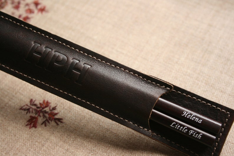 May include: A brown leather case with the initials "HPH" embossed on the front. The case contains two wooden chopsticks with the names "Helena" and "Little Fish" engraved on them.