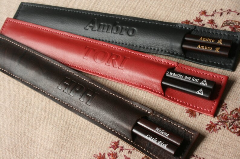 May include: Three leather chopsticks cases with personalized names. The cases are black, red, and brown. The chopsticks are visible inside the cases.