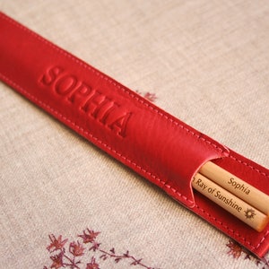 May include: Red leather chopsticks case with the name "Sophia" embossed on the front. The case contains two wooden chopsticks with the text "Sophia Ray of Sunshine" and a sun symbol engraved on them.