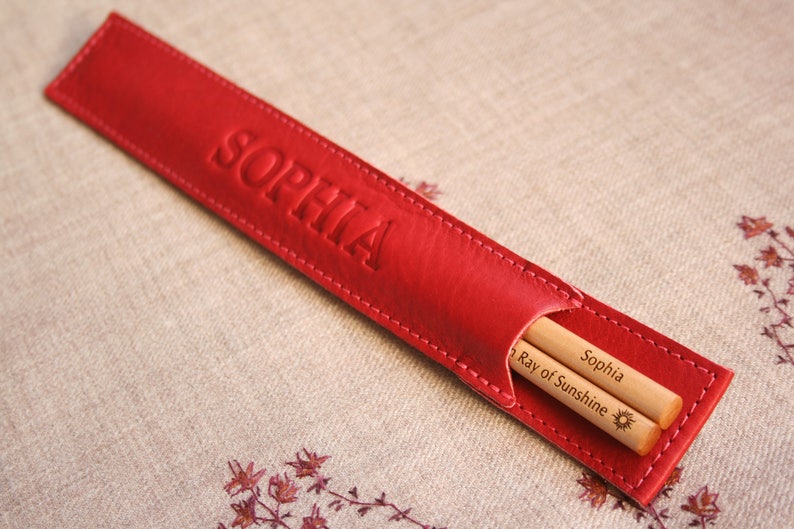 May include: Red leather chopsticks case with the name "Sophia" embossed on the front. The case contains two wooden chopsticks with the text "Sophia Ray of Sunshine" and a sun symbol engraved on them.