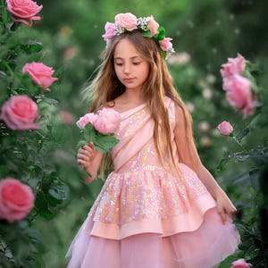 May include: A young person wearing a pink dress with a sequined bodice and layered skirt, holding pink roses. They are also wearing a floral crown. The background is a garden with pink roses.