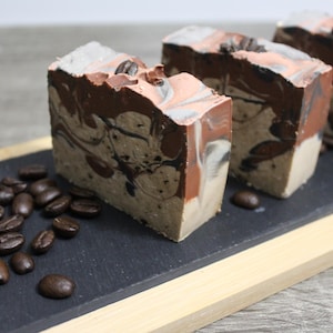 Könnte beinhalten: Drei handgemachte Seifenstücke mit einem Kaffee- und Braunstirn-Design. Die Seifenstücke sind auf einer schwarzen Schieferplatte mit Kaffeekörnern drumherum angeordnet.