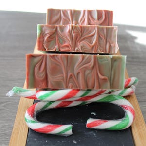 May include: Three bars of handmade soap stacked on top of each other. The soap is a swirl of pink and green. The soap is sitting on a wooden surface with candy canes.