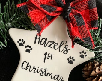May include: White ceramic star ornament with black paw prints and the text "Hazel's 1st Christmas". The ornament is decorated with a red and black buffalo plaid bow.