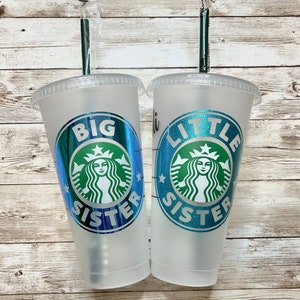 May include: Two clear plastic cups with lids and straws. The cups have a green and blue design with a mermaid and the words "Big Sister" and "Little Sister".
