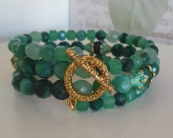 May include: Three green beaded bracelets stacked together. The beads are in shades of green, with faceted and round shapes. A gold-coloured clasp fastens the bracelets, with decorative gold and teal accents.