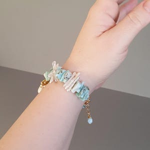 Puede incluir: Un brazalete hecho a mano con piedras preciosas azul claro y blancas, una cadena dorada y un peque&ntilde;o dije azul. El brazalete est&aacute; adornado con piedras y perlas de forma irregular, creando un accesorio &uacute;nico y elegante.