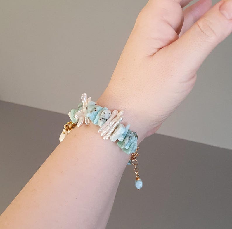 Puede incluir: Un brazalete hecho a mano con piedras preciosas azul claro y blancas, una cadena dorada y un peque&ntilde;o dije azul. El brazalete est&aacute; adornado con piedras y perlas de forma irregular, creando un accesorio &uacute;nico y elegante.