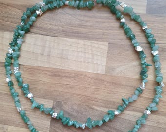 May include: A double-strand necklace composed of small, irregular green gemstone beads. The necklace is accented with silver-coloured elements and rests on a wooden surface. The gemstones have a natural, organic appearance.