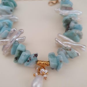 Puede incluir: Un brazalete hecho a mano con piedras de larimar azul claro y perlas de agua dulce blancas. El brazalete tiene una cadena dorada y un dije dorado con un gran colgante de perla en forma de l&aacute;grima.