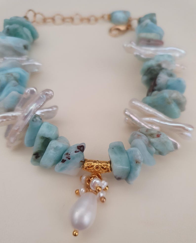 Puede incluir: Un brazalete hecho a mano con piedras de larimar azul claro y perlas de agua dulce blancas. El brazalete tiene una cadena dorada y un dije dorado con un gran colgante de perla en forma de l&aacute;grima.