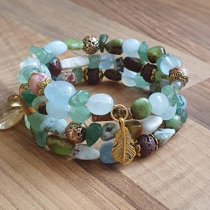May include: Three beaded bracelets with a mix of green, blue, brown, and white natural stone beads. The bracelets have gold-coloured details and a gold feather charm. The bracelets are displayed on a wooden surface.