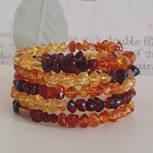 Könnte beinhalten: Ein Stapel Bernsteinarmbänder. Die Armbänder bestehen aus kleinen, unregelmäßig geformten Perlen in Gelb-, Orange- und Dunkelrottönen. Die Armbänder sind umeinander gewickelt und erzeugen einen geschichteten Effekt. Der Hintergrund ist unscharf.