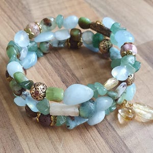 May include: A wrap bracelet with a mix of light blue, green, and brown gemstone beads. The bracelet has gold-coloured accents and a dangling charm with a teardrop-shaped stone. The bracelet is displayed on a wooden surface.