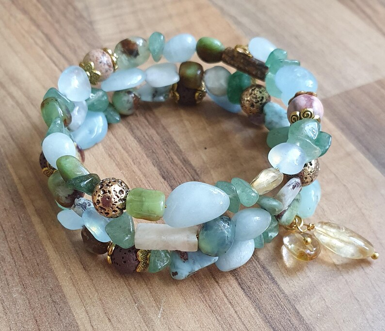 May include: A wrap bracelet with a mix of light blue, green, and brown gemstone beads. The bracelet has gold-coloured accents and a dangling charm with a teardrop-shaped stone. The bracelet is displayed on a wooden surface.