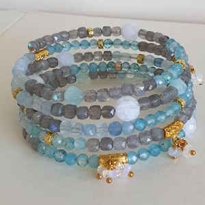 May include: A multi-strand wrap bracelet with blue, grey, and white beads. The bracelet has gold-coloured accents and small dangling charms. The beads are various shapes and sizes, creating a layered effect.