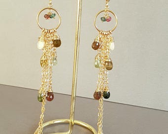 May include: A pair of gold-tone dangle earrings featuring a circular hoop at the top with small multi-coloured gemstone beads. Below the hoop, multiple gold chains hang down, each accented with small, teardrop-shaped, multi-coloured tourmaline beads.