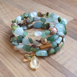 May include: Three gemstone bracelets stacked together, featuring green, blue, and brown beads. The bracelets have gold-coloured accents and a clear teardrop charm. Displayed on a wooden surface.