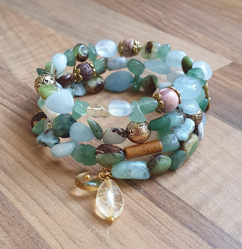 May include: Three gemstone bracelets stacked together, featuring green, blue, and brown beads. The bracelets have gold-coloured accents and a clear teardrop charm. Displayed on a wooden surface.