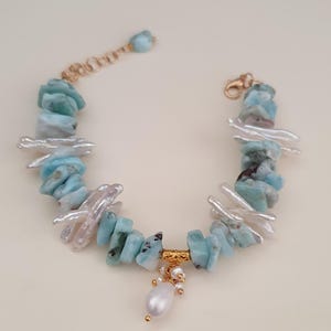 Puede incluir: Una pulsera hecha a mano con piedras de larimar azul claro y perlas de agua dulce blancas. La pulsera tiene un cierre dorado y una barra decorativa dorada con una perla colgante y peque&ntilde;as cuentas doradas. La pulsera mide aproximadamente 18 cm de largo.
