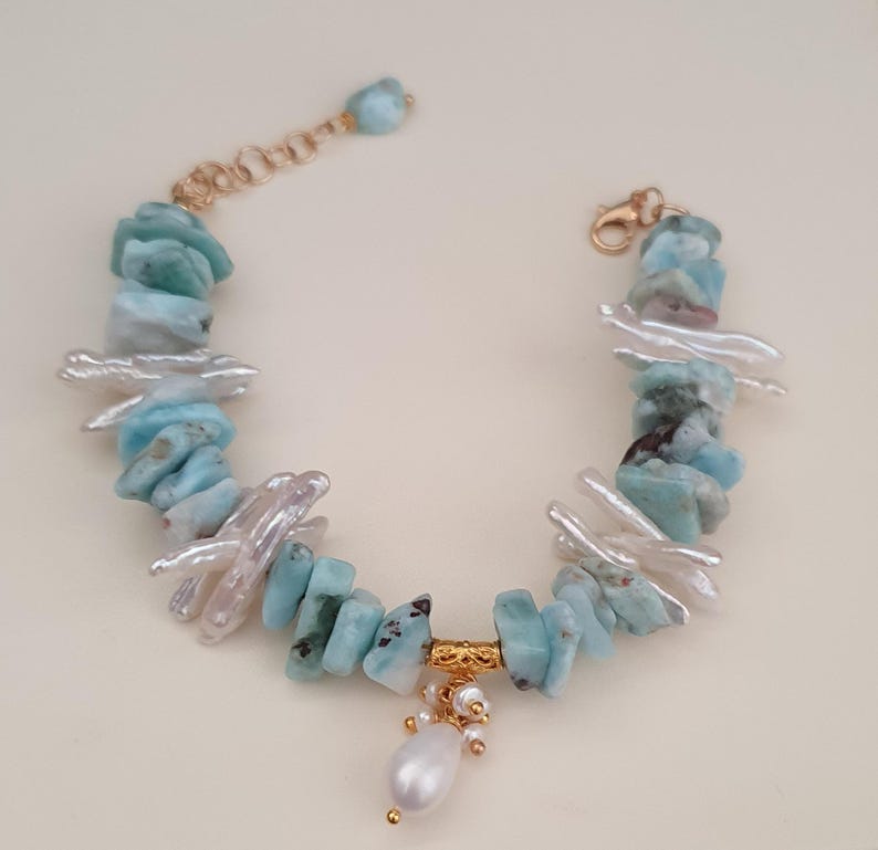 Puede incluir: Una pulsera hecha a mano con piedras de larimar azul claro y perlas de agua dulce blancas. La pulsera tiene un cierre dorado y una barra decorativa dorada con una perla colgante y peque&ntilde;as cuentas doradas. La pulsera mide aproximadamente 18 cm de largo.