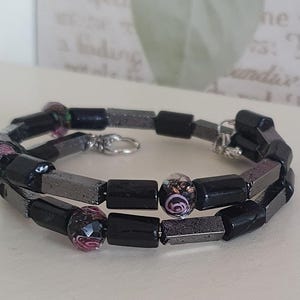 Op de afbeelding: Een wikkelarmband met zwarte en grijze rechthoekige kralen, afgewisseld met decoratieve glazen kralen in roze, paars en groen. De armband is vastgezet met een zilverkleurige sluiting.