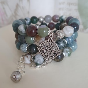 Può includere: Un braccialetto multifilo realizzato con perline di agata verde, marrone e bianca con un ciondolo d'argento. Il ciondolo ha una forma quadrata con un motivo floreale.