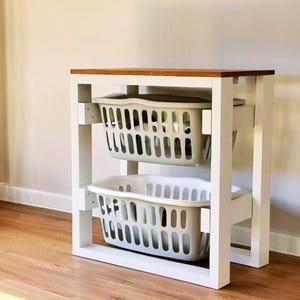 Può includere: Un divisore per bucato in legno bianco con due cesti per bucato in plastica grigia rimovibili. La parte superiore è in legno tinto. Il divisore è su un pavimento in legno.