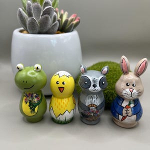 May include: Four painted wooden figurines: a green frog with flowers, a yellow chick hatching from an egg, a grey raccoon with a basket, and a brown rabbit with a carrot. The figurines are detailed and stand upright.